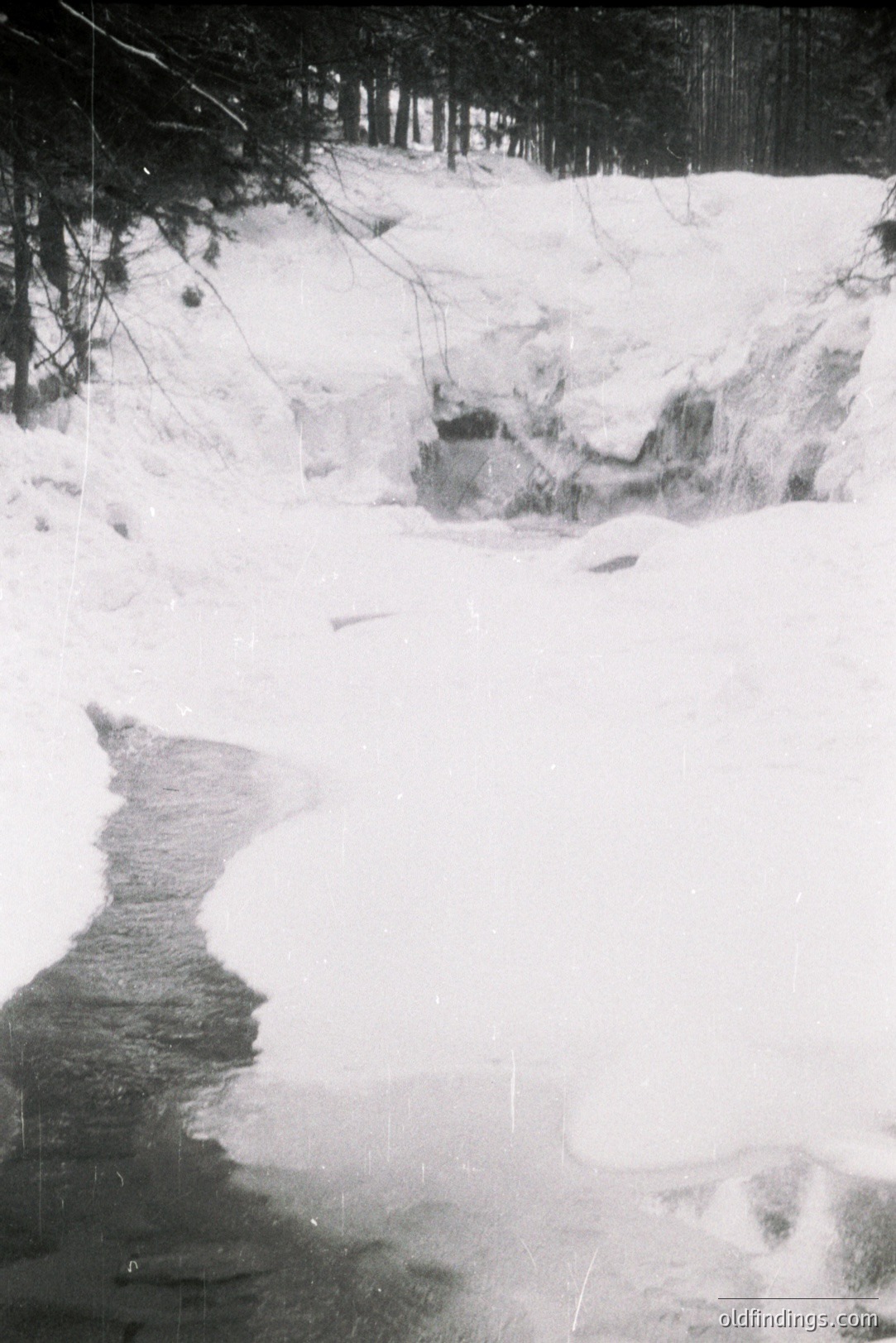 A frozen stream flows through a snow-covered landscape with a dense treeline in the background. The ice surface shows subtle texture. Likely a rural, winter scene, possibly captured in the mid-20th century due to the film grain and aesthetic.