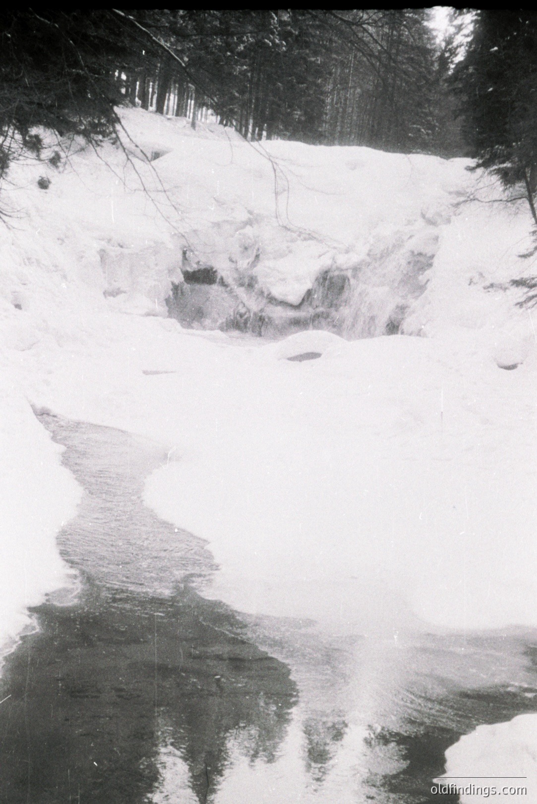 Monochrome landscape depicting a partially frozen stream flowing through a snow-covered forest. The water reflects dark tree branches and surrounding foliage. Appears to be a remote, natural environment. Likely mid-20th century based on photographic style. Possible stock photography reference for winter scenes.
