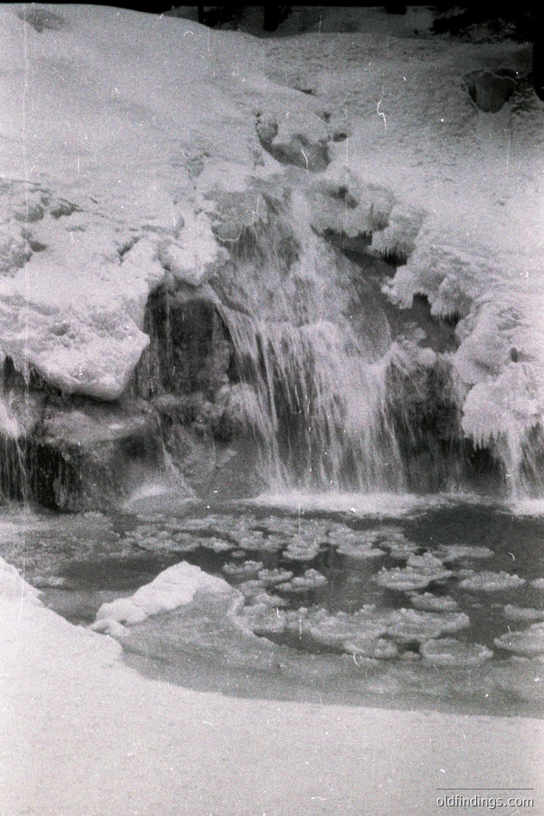A frozen waterfall cascades over ice formations in a dramatic, monochrome landscape. Water droplets hang suspended, captured mid-air. The scene evokes a sense of icy wilderness. Likely a 20th-century photographic study of a natural phenomenon. Excellent potential for design and research use.