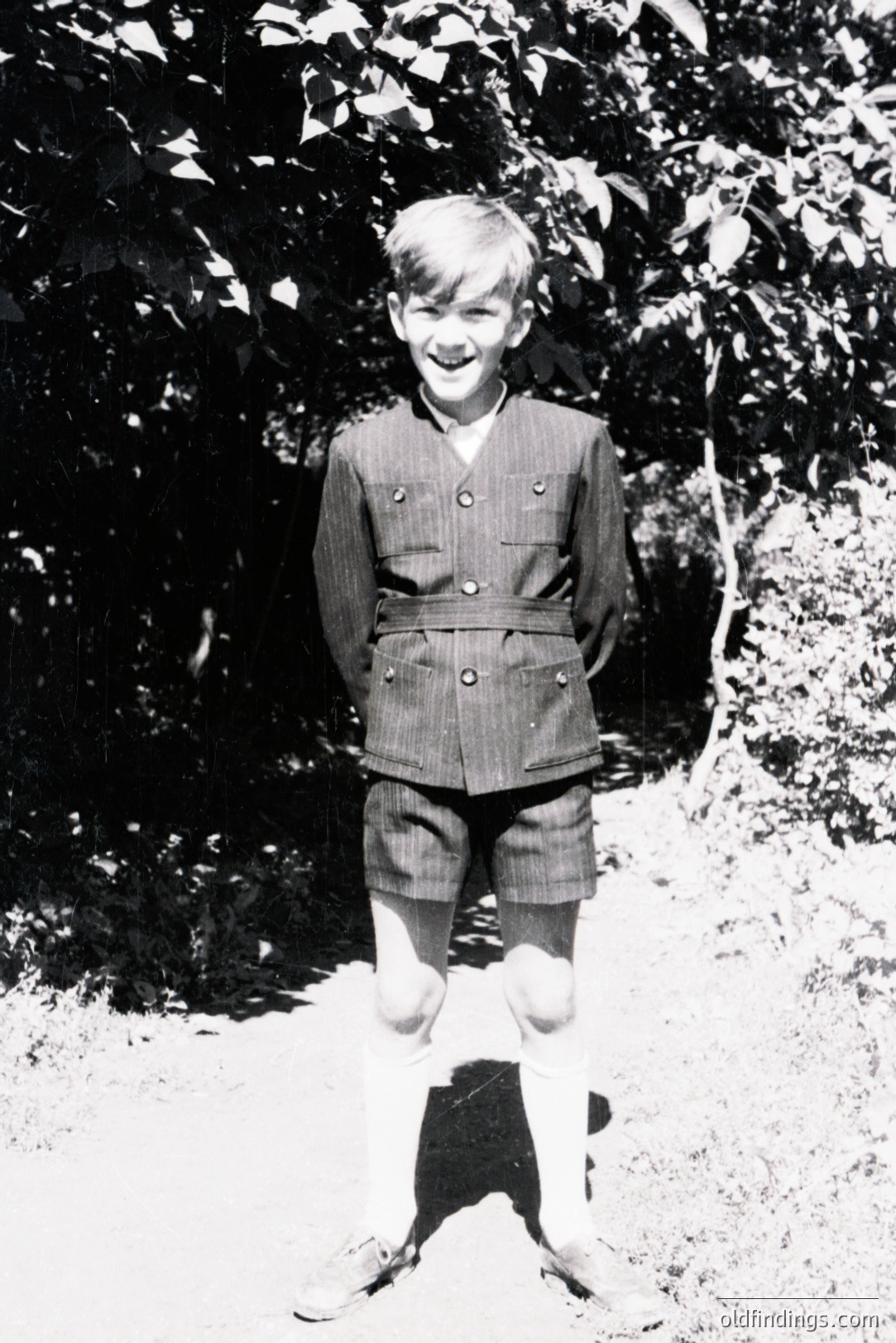 Young boy in a tailored double-breasted jacket & shorts, knee socks, stands outdoors. Appears to be a formal school or uniform attire. Likely 1960s or 70s, based on style. Background features dense foliage and a textured path. Classic portrait style, potential for vintage design reference.
