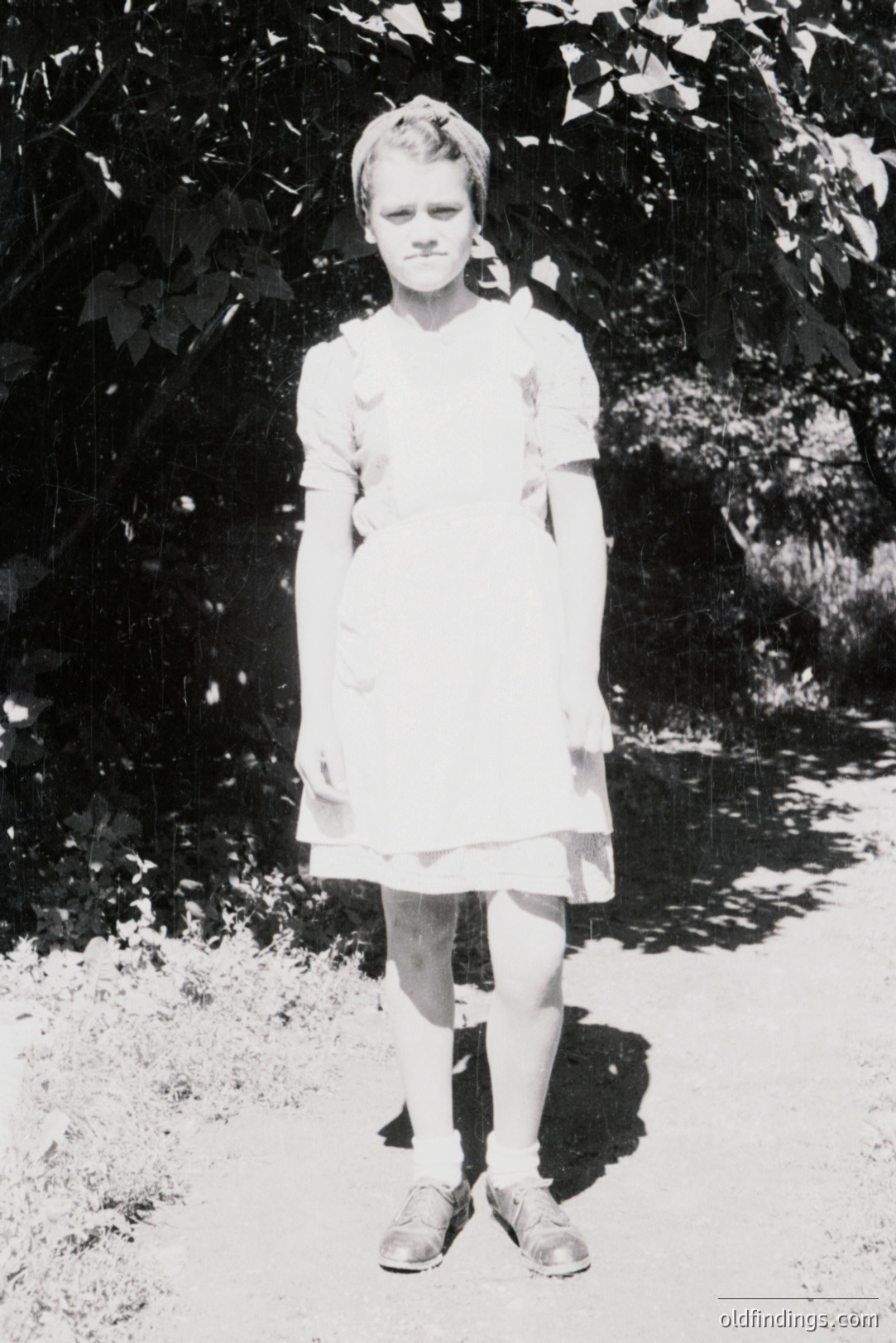 A young girl stands on a stone path, wearing a simple, knee-length dress and dark shoes with socks. Her hair is styled in short waves. The backdrop consists of dense foliage, suggesting a garden or wooded area. Likely a studio portrait, circa 1930s-1950s. Valuable for family history and vintage fashion references.