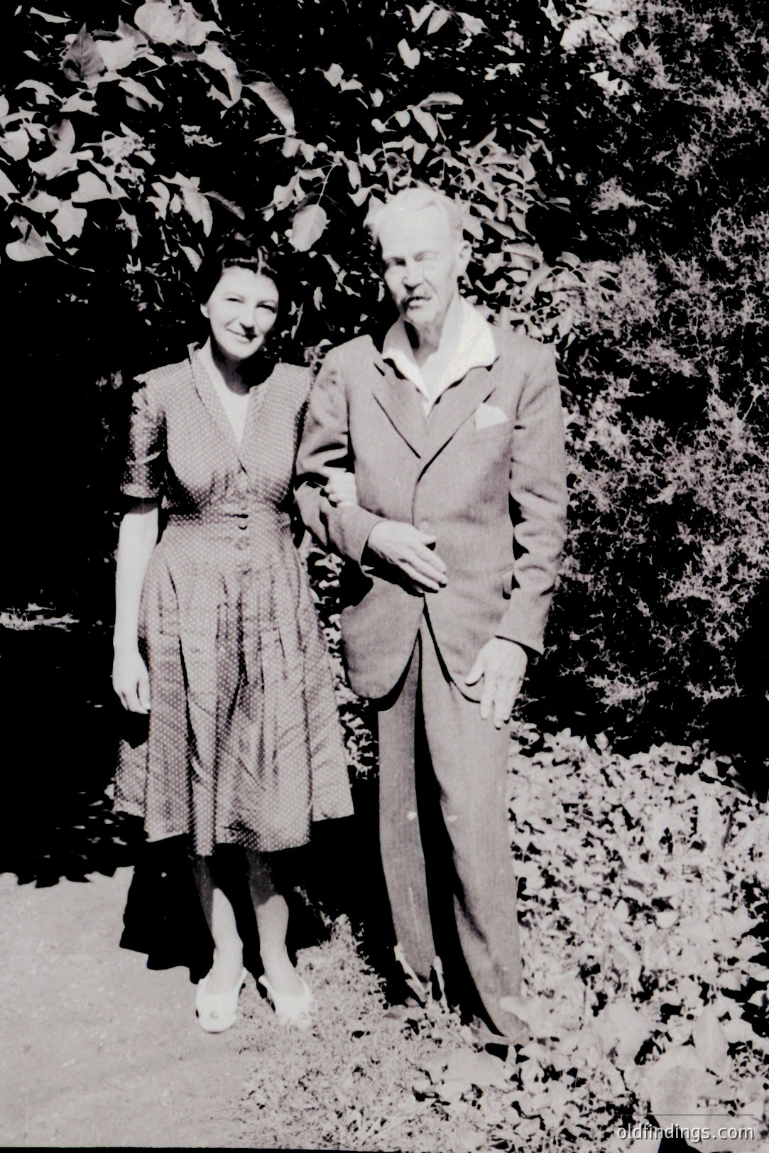 A posed portrait of a young woman and older gentleman, likely a couple, standing outdoors among foliage. Woman wears a knee-length, printed dress; the man, a tailored suit. Style suggests a 1940s or 1950s setting. Likely a family or personal snapshot. Good for historical costuming references.