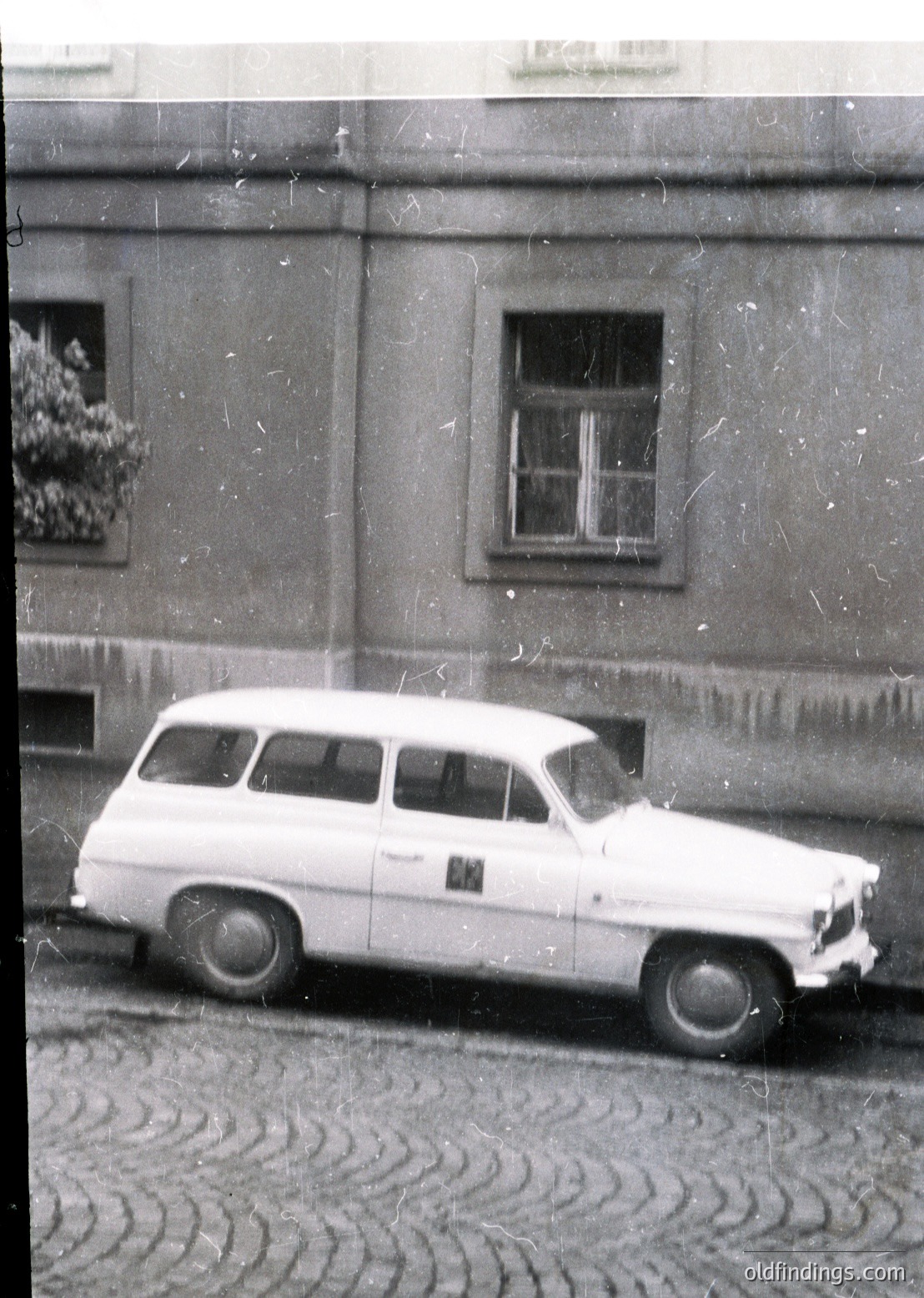 Vintage white station wagon parked on cobblestone street beside Soviet-era building with floral window box. Classic 1960s Eastern Bloc design, likely Eastern Europe.