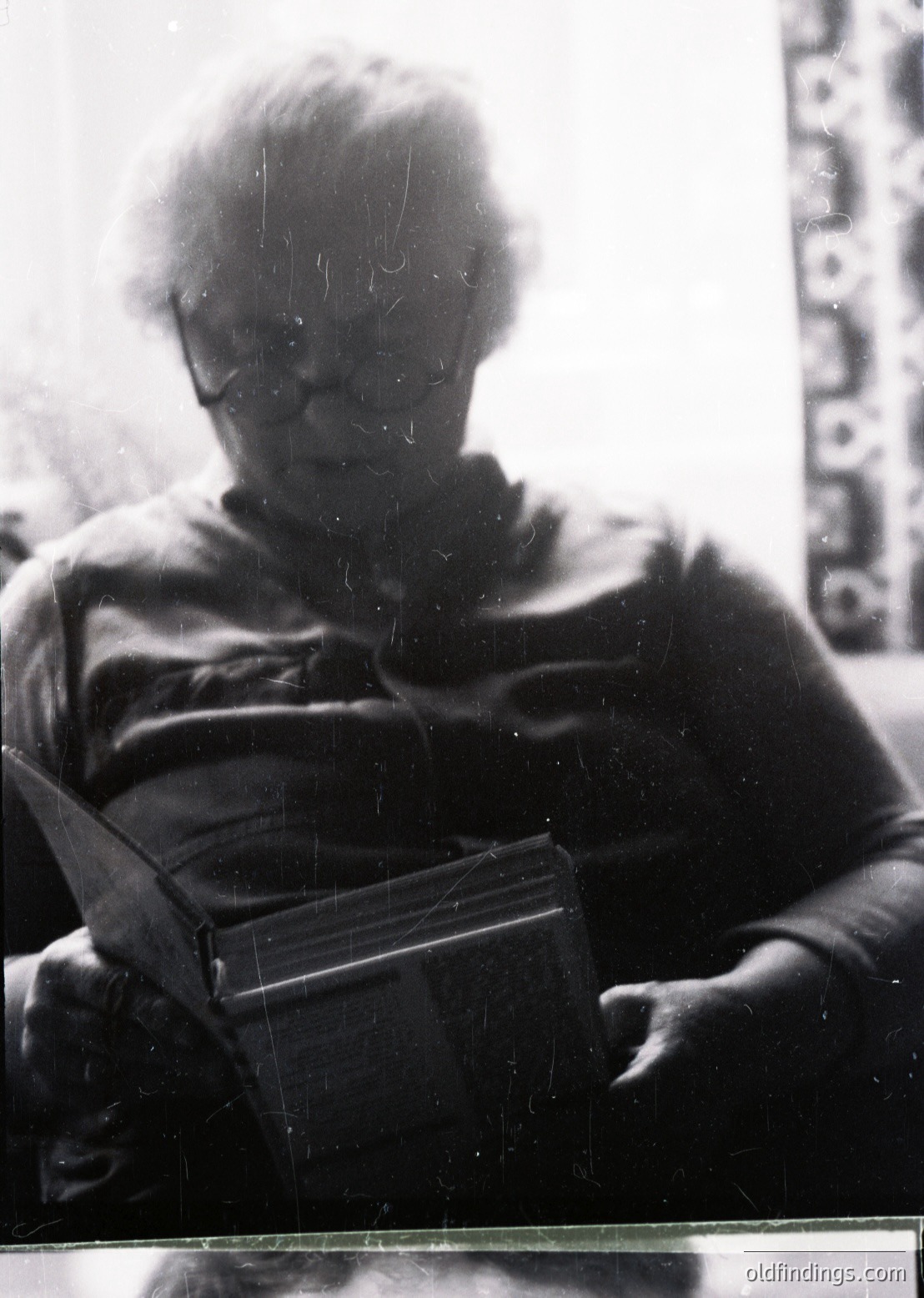 Vintage black-and-white portrait of an individual holding a vintage camera, likely a Leica-style rangefinder. The person’s short, textured hair and round glasses suggest mid-20th century fashion. Background shows patterned wallpaper with floral motifs, hinting at mid-century domestic interior. Potential photography scene.