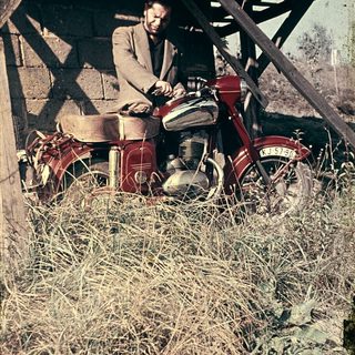 Man sitting on vintage red motorcycle in dry grass.