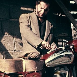 Man sitting on vintage red motorcycle in dry grass.