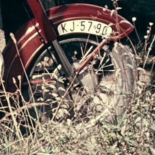 Man sitting on vintage red motorcycle in dry grass.