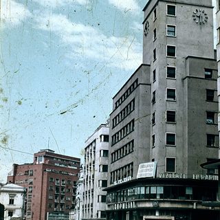 Mid-century urban street scene with tall buildings and pharmacy sign.