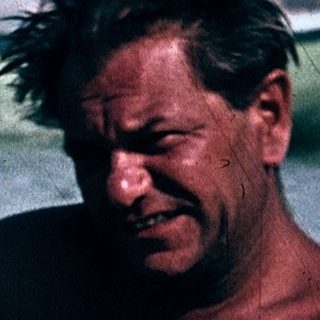 Shirtless man sitting in shallow ocean water.