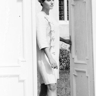 Woman in 1960s-style dress and hat near open door.