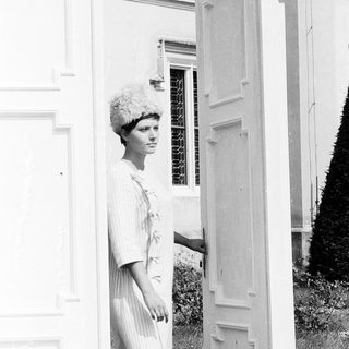 Woman in 1960s-style dress and hat near open door.