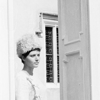 Woman in 1960s-style dress and hat near open door.