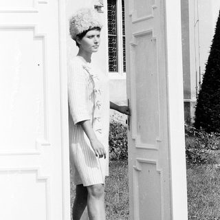 Woman in 1960s-style dress and hat near open door.