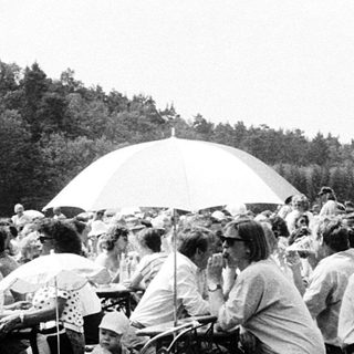Outdoor gathering with people seated at tables under umbrellas.