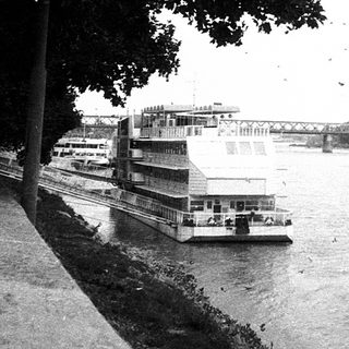 River scene with ferry, bridge, and tree-lined embankment.