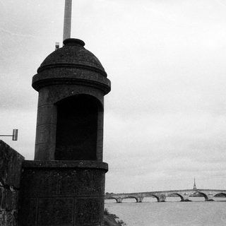 Stone sentry box by river with bridge and city skyline.