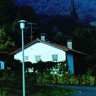 Rural house with sloped roof, surrounded by greenery and hills.