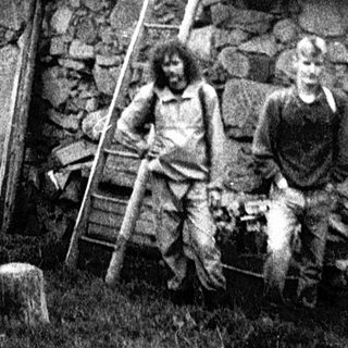 Rural scene with two men near rustic stone house and wooden ladder.