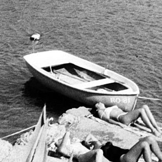A man stands by a small boat on rocky shore near calm waters.