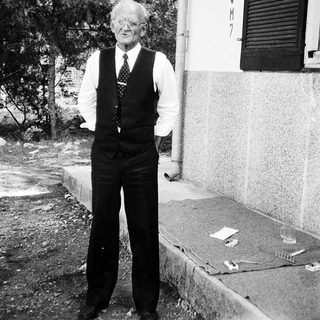 Elderly man standing outdoors in vintage vest and tie.