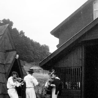 Three musicians playing traditional instruments outdoors near rustic buildings.