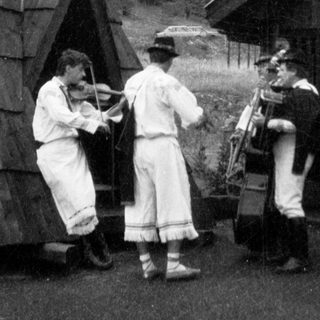 Three musicians playing traditional instruments outdoors near rustic buildings.