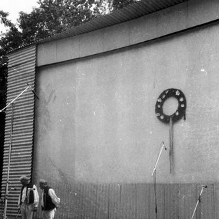 Two men standing near a wreath on a plain wall.
