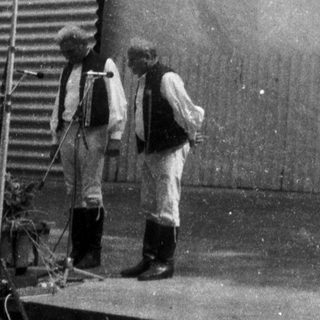 Two men standing near a wreath on a plain wall.