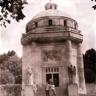 Neoclassical mausoleum with statues, three men standing outside.