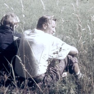 Two people sitting in tall grass overlooking rural landscape.