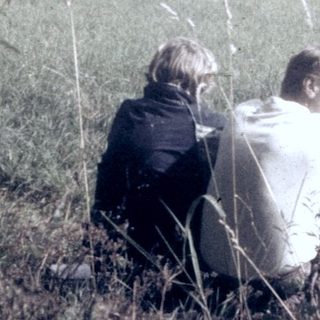 Two people sitting in tall grass overlooking rural landscape.