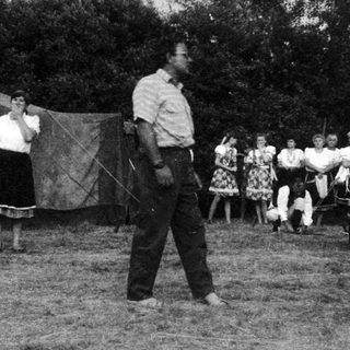 Camp setting with man demonstrating archery to onlookers.