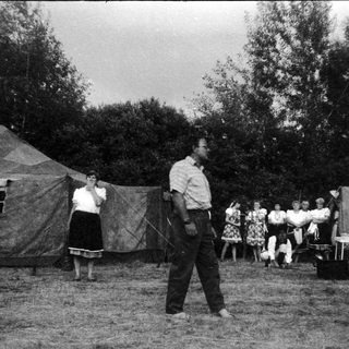Camp setting with man demonstrating archery to onlookers.