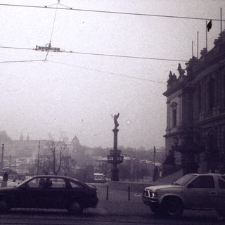 Historic city street with tram wires, cars, and castle in foggy background.
