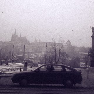Historic city street with tram wires, cars, and castle in foggy background.
