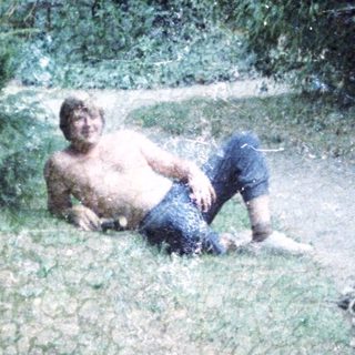 Shirtless man sitting wet in forested area.