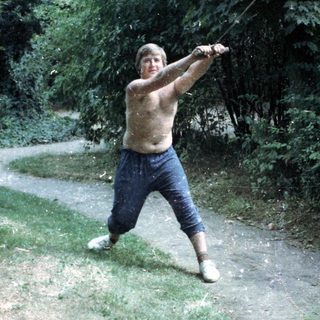 Shirtless man throwing a long pole outdoors.