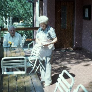 Two elderly people sitting and standing outdoors on a rainy day.