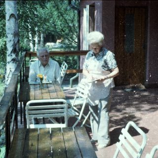 Two elderly people sitting and standing outdoors on a rainy day.