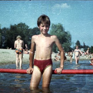 Shirtless boy in red swim trunks at crowded pool.