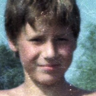 Shirtless boy in red swim trunks at crowded pool.