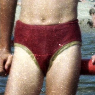 Shirtless boy in red swim trunks at crowded pool.