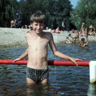 Shirtless boy standing in shallow water at crowded beach.