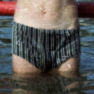 Shirtless boy standing in shallow water at crowded beach.