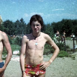 Two shirtless young men posing outdoors by water.