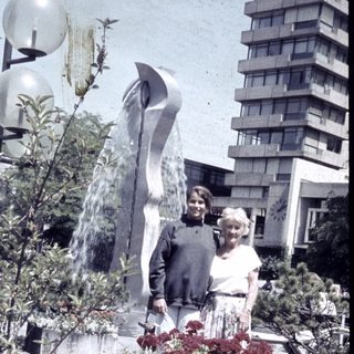 Two people posing beside modern fountain sculpture and blooming flowers.