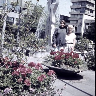 Two people posing beside modern fountain sculpture and blooming flowers.