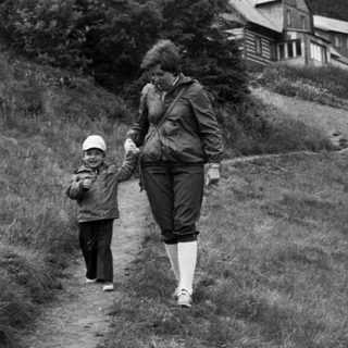 Adult leading child on grassy hillside near forest cabin.
