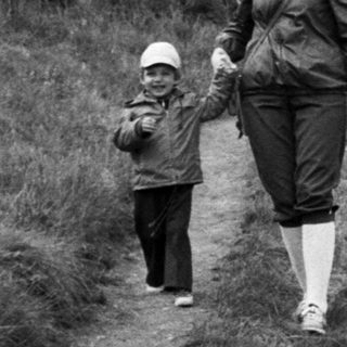 Adult leading child on grassy hillside near forest cabin.