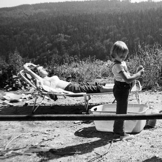 Child playing near a tub outdoors with relaxed adult in background.